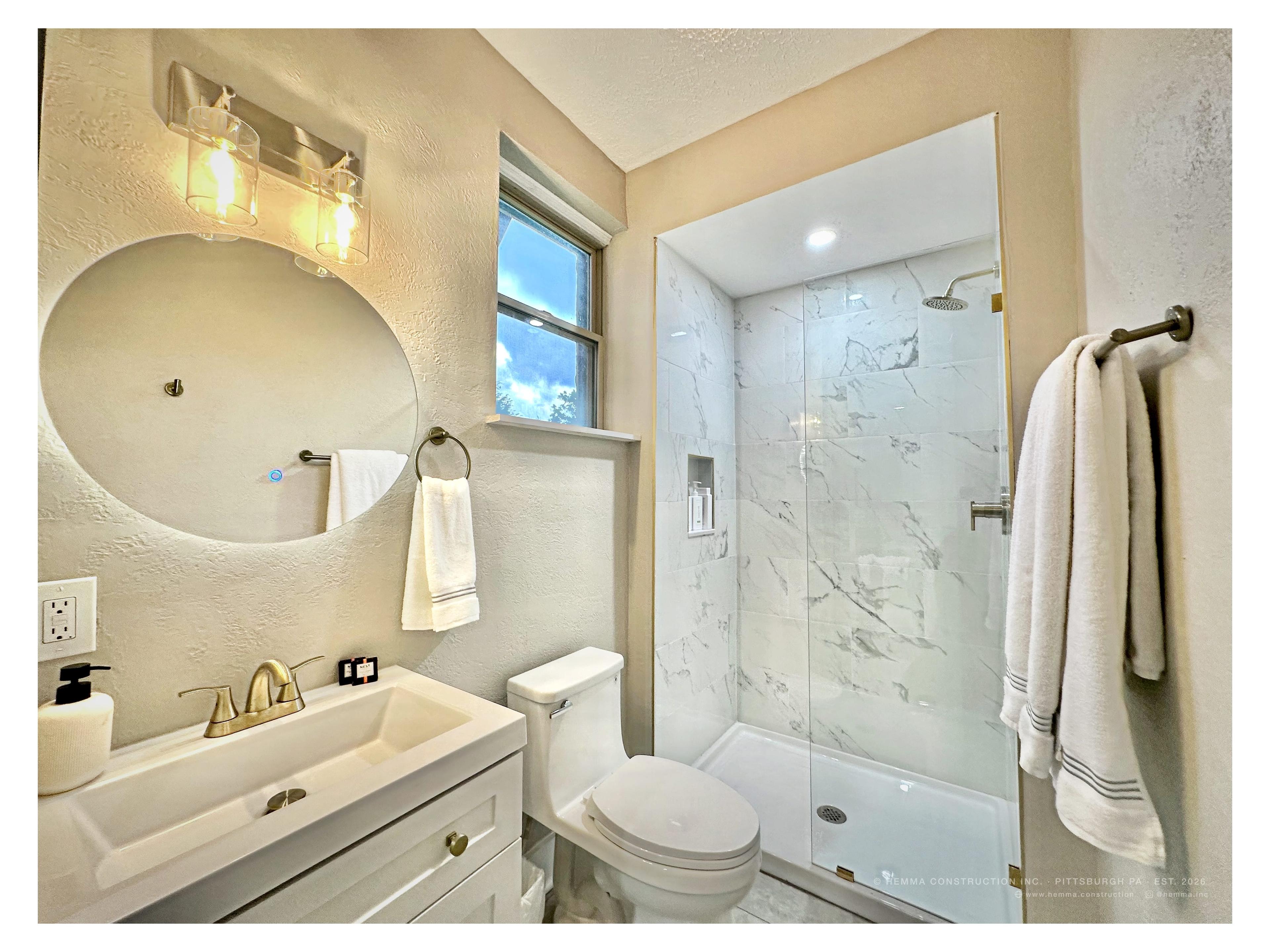 Bathroom — backlit round mirror, gold faucet, marble walk-in shower